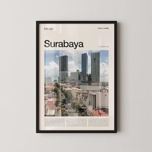Op de afbeelding: Een ingelijste print van Surabaya, Indonesië, met een stadsgezicht met hoge gebouwen onder een bewolkte hemel. Het woord "Surabaya" staat prominent afgebeeld, met "Indonesië" eronder. De print zit in een zwarte lijst.
