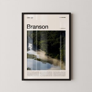 May include: Framed art print featuring the word "Branson" in bold black letters above a landscape photograph. The photograph shows a river winding through a forest, with mist rising from the water. The print is in a black frame.