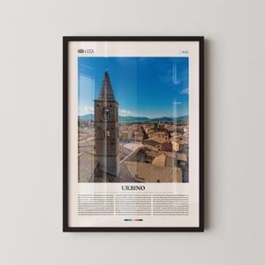 May include: Framed print featuring a detailed view of Urbino, Italy, showcasing a tall bell tower and cityscape under a bright blue sky. The print includes the text "URBINO" and additional descriptive text.