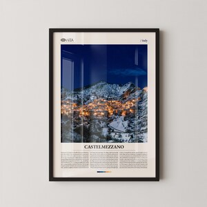 Könnte beinhalten: Ein gerahmter Druck von Castelmezzano, Italien, bei Nacht. Das Bild zeigt einen schneebedeckten Berg mit einem Dorf, das von warmen Lichtern beleuchtet wird. Der Text "CASTELMEZZANO" befindet sich unten, mit zusätzlichem Text darunter. Der Rahmen ist schwarz.