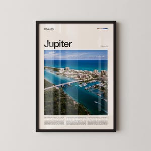 Könnte beinhalten: Ein gerahmter Druck mit einer Luftaufnahme von Jupiter, Florida, mit dem Wort "Jupiter". Das Bild zeigt eine Brücke, Boote, Gebäude und den Ozean unter blauem Himmel. Der Rahmen ist schwarz.