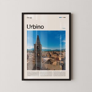 May include: Framed print featuring a photograph of Urbino, Italy, with a tall bell tower dominating the skyline. The image is set against a bright blue sky, with the word "Urbino" in large black letters at the top. The frame is black.