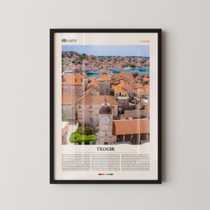 Puede incluir: Impresión enmarcada que presenta una vista panorámica de Trogir, Croacia, con tejados de tejas rojas y una torre del reloj. La imagen incluye el texto "Trogir" y "/ Croacia". La impresión está en un marco negro y se muestra contra una pared blanca.