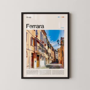 May include: A framed print depicting a street scene in Ferrara, Italy. The image shows a narrow street with buildings in yellow and orange, under a blue sky. The word "Ferrara" is displayed at the top of the print.