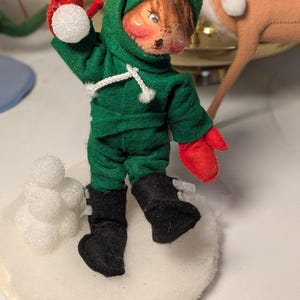 May include: A felt doll dressed in a green hooded outfit with red mittens and black boots, throwing a white foam snowball. The doll stands on a circular white base next to a small stack of white foam snowballs, with a painted face and brown hair.