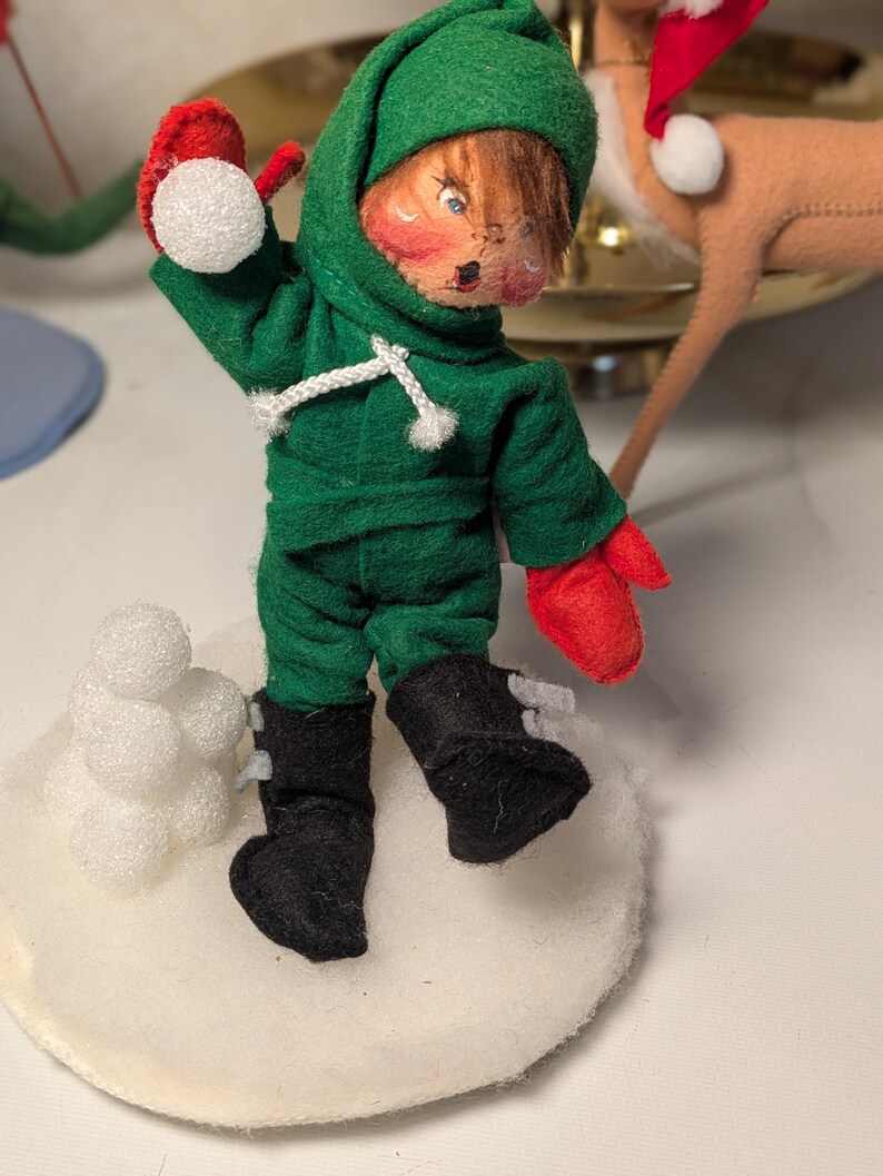 May include: A felt doll dressed in a green hooded outfit with red mittens and black boots, throwing a white foam snowball. The doll stands on a circular white base next to a small stack of white foam snowballs, with a painted face and brown hair.