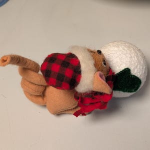 May include: A tan felt mouse figurine wearing a red and black buffalo plaid hat and scarf, featuring a long curled tail and holding a textured white snowball with a small green felt leaf attached to the side of the round white sphere.