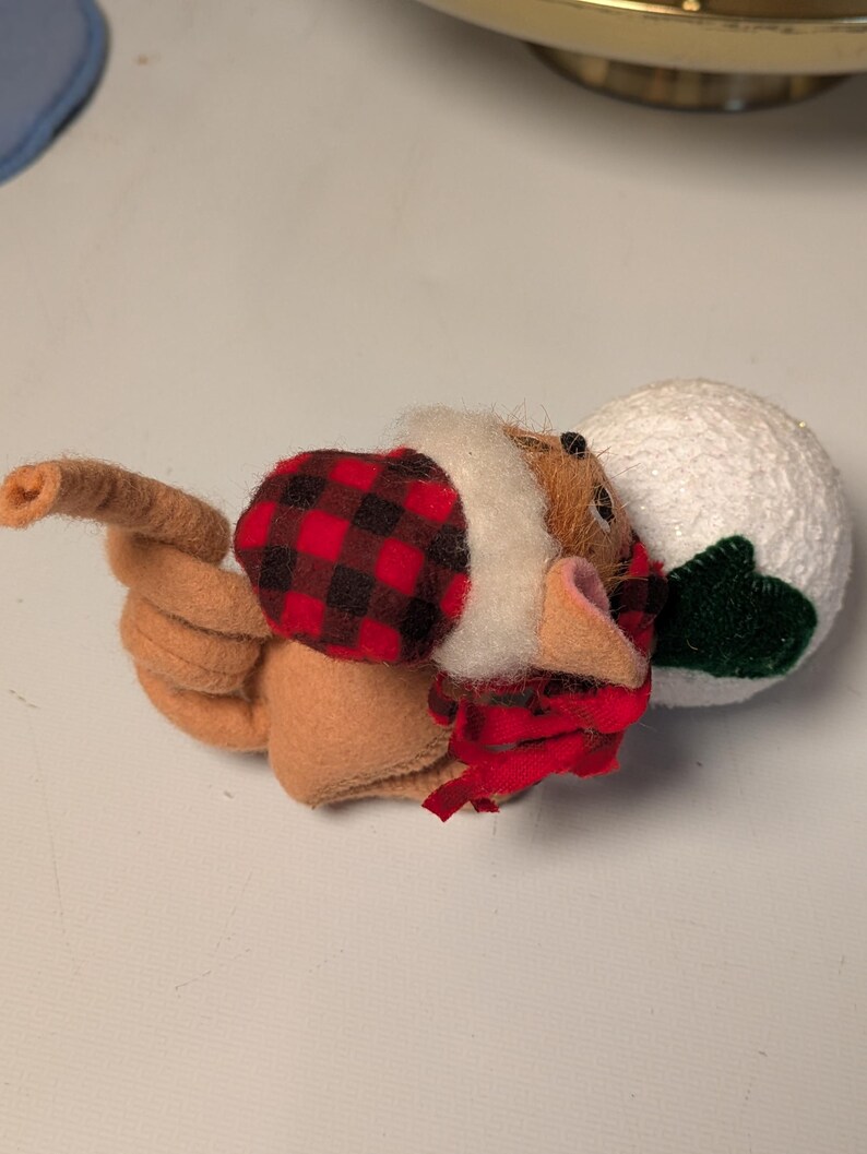 May include: A tan felt mouse figurine wearing a red and black buffalo plaid hat and scarf, featuring a long curled tail and holding a textured white snowball with a small green felt leaf attached to the side of the round white sphere.