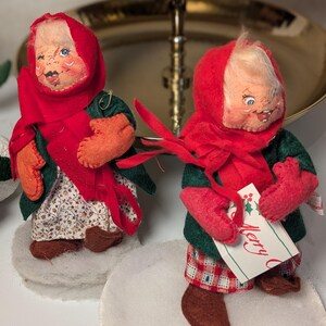 May include: Two fabric dolls with painted faces, wearing red headscarves, dark green coats, and red mittens. One doll wears a floral skirt, the other red plaid pants. They stand on white felt circles. One doll holds a tag with the text Merry C.