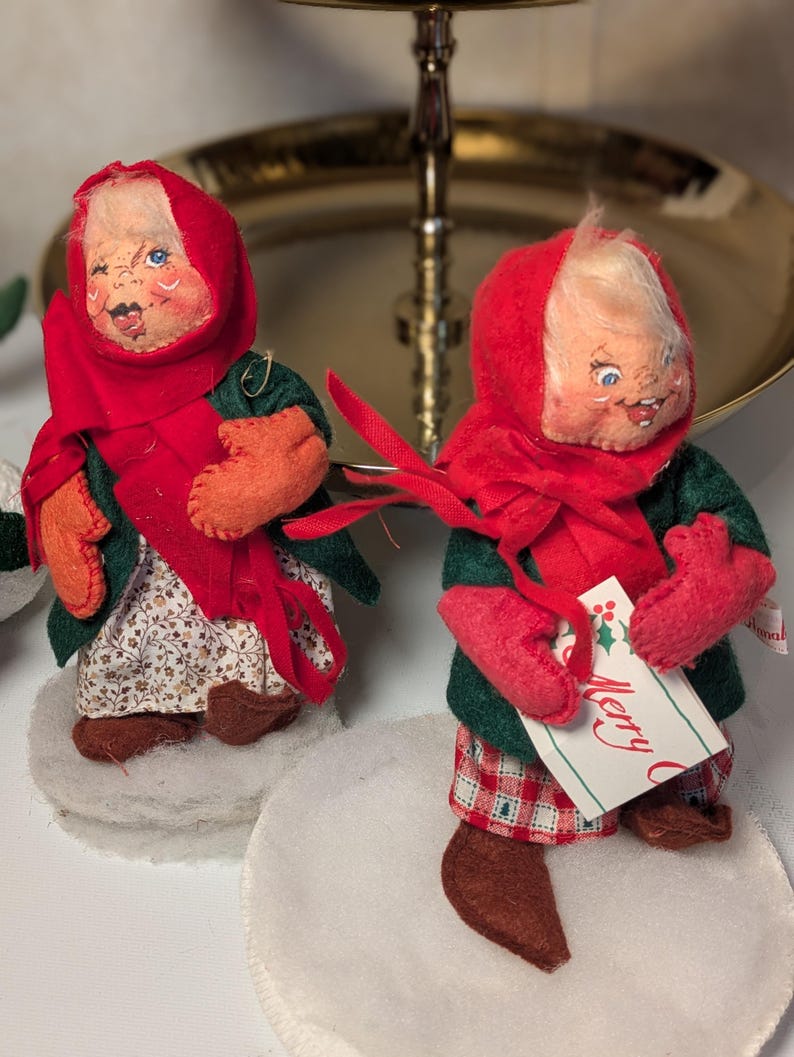 May include: Two fabric dolls with painted faces, wearing red headscarves, dark green coats, and red mittens. One doll wears a floral skirt, the other red plaid pants. They stand on white felt circles. One doll holds a tag with the text Merry C.