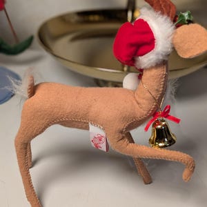 May include: A tan felt reindeer ornament featuring a red and white Santa hat, a small gold-colored bell hanging from a thin gold chain around its neck, and a red ribbon bow. The reindeer has a white tail and a small white tag on its side.