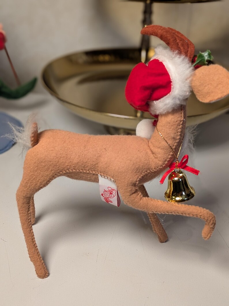 May include: A tan felt reindeer ornament featuring a red and white Santa hat, a small gold-colored bell hanging from a thin gold chain around its neck, and a red ribbon bow. The reindeer has a white tail and a small white tag on its side.