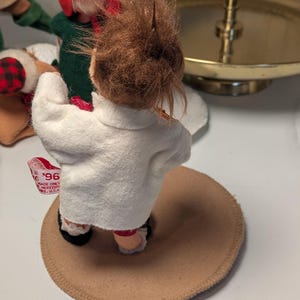 May include: A small doll with brown hair wearing a white felt jacket, standing on a circular tan base. A white fabric tag attached to the jacket features red text that reads '96' and 'MADE ONLY'. The doll is positioned near other figures.