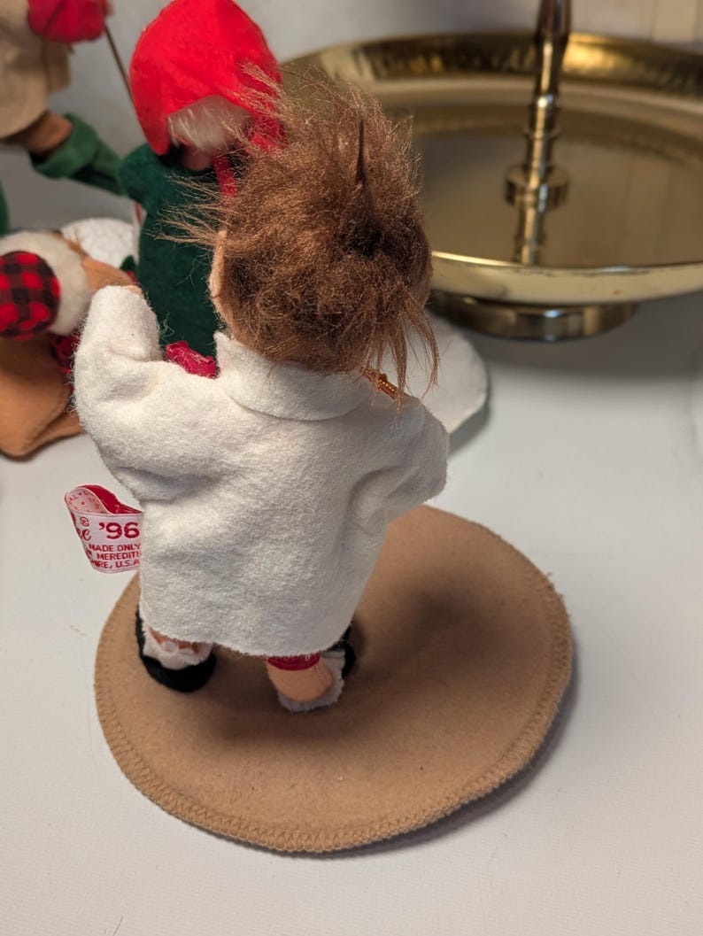 May include: A small doll with brown hair wearing a white felt jacket, standing on a circular tan base. A white fabric tag attached to the jacket features red text that reads '96' and 'MADE ONLY'. The doll is positioned near other figures.