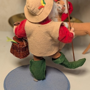 May include: A felt figurine wearing a tan hat with small red, yellow, and green accents, a tan vest over a red long-sleeved shirt, and green boots. The figure carries a brown woven basket and a thin wooden fishing rod, standing on a blue base.