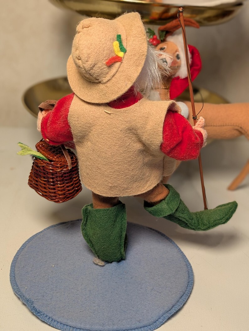 May include: A felt figurine wearing a tan hat with small red, yellow, and green accents, a tan vest over a red long-sleeved shirt, and green boots. The figure carries a brown woven basket and a thin wooden fishing rod, standing on a blue base.