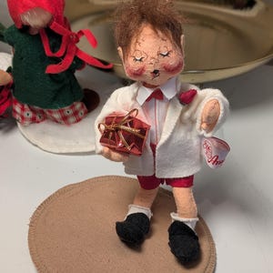 May include: A fabric doll with brown hair, wearing a white jacket, red shorts, and black shoes, holding a small red gift box with gold ribbon. The doll has a white tag on the sleeve with red script text and stands on a circular tan base.