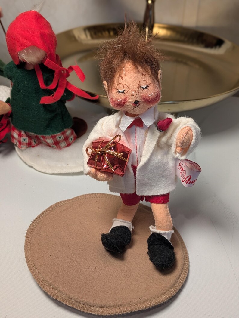 May include: A fabric doll with brown hair, wearing a white jacket, red shorts, and black shoes, holding a small red gift box with gold ribbon. The doll has a white tag on the sleeve with red script text and stands on a circular tan base.