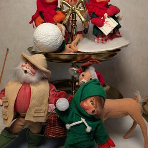 May include: A two-tiered gold metal stand holding three felt dolls with painted faces, red scarves, and winter clothing. Below, a bearded doll in a tan vest and hat stands beside a green-hooded doll holding a snowball, with a felt deer nearby.