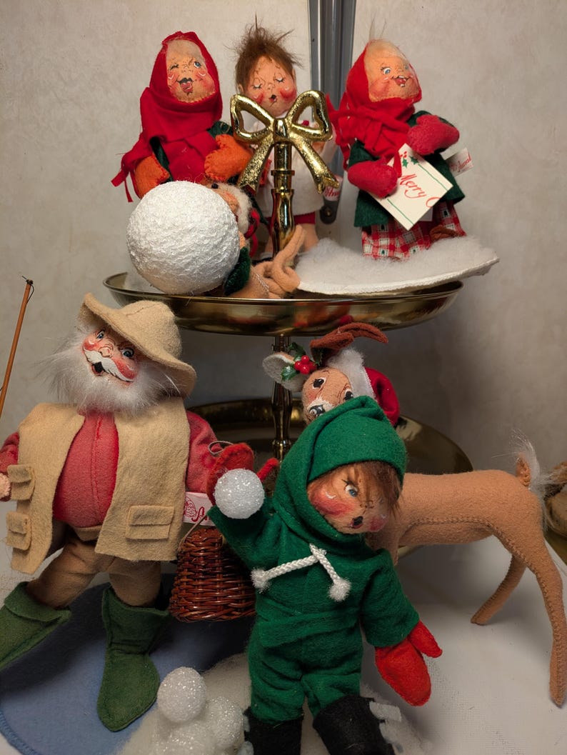 May include: A two-tiered gold metal stand holding three felt dolls with painted faces, red scarves, and winter clothing. Below, a bearded doll in a tan vest and hat stands beside a green-hooded doll holding a snowball, with a felt deer nearby.