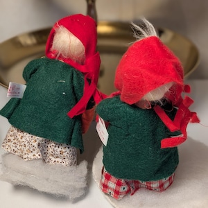 May include: Two small figurines wearing red felt hoods and dark green felt coats. One figurine wears a white skirt with a brown floral pattern, while the other wears red and white checkered pants. Both have white hair and small white tags.