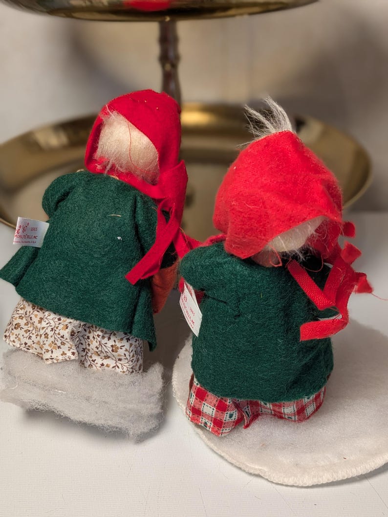 May include: Two small figurines wearing red felt hoods and dark green felt coats. One figurine wears a white skirt with a brown floral pattern, while the other wears red and white checkered pants. Both have white hair and small white tags.