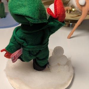 May include: A felt figurine dressed in a green hooded robe with red mittens, standing on a white circular base with a small pile of white glittery spheres. A pink fabric tag attached to the side of the robe displays the text '96'.