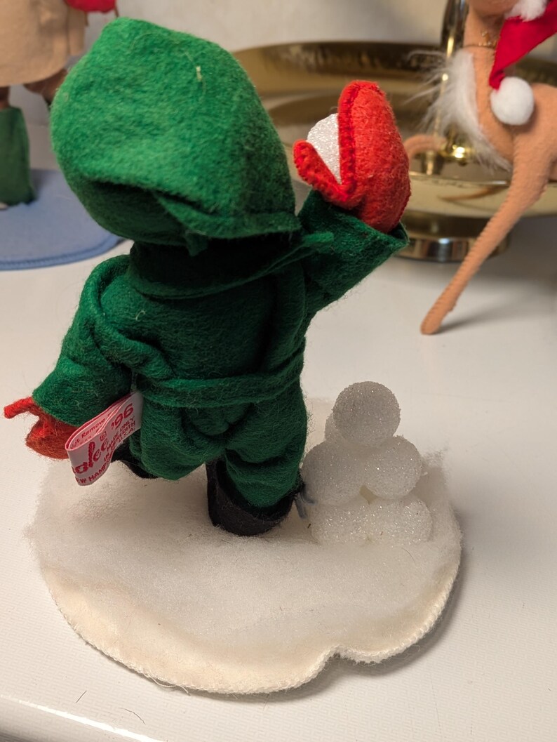 May include: A felt figurine dressed in a green hooded robe with red mittens, standing on a white circular base with a small pile of white glittery spheres. A pink fabric tag attached to the side of the robe displays the text '96'.