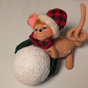 May include: A tan felt mouse figurine wearing a red and black buffalo plaid hat with a white faux fur trim and a matching scarf. The mouse has dark green mittens and is holding a large, textured, glittery white snowball in its front paws.