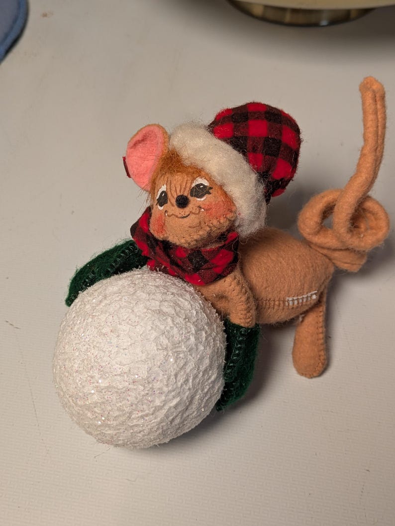 May include: A tan felt mouse figurine wearing a red and black buffalo plaid hat with a white faux fur trim and a matching scarf. The mouse has dark green mittens and is holding a large, textured, glittery white snowball in its front paws.