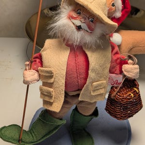 May include: A figurine of a bearded figure wearing a tan hat, a red shirt, a tan vest, and green boots. The figure holds a thin brown fishing rod in one hand and a small brown woven basket in the other. A white tag with red text is attached to the basket.