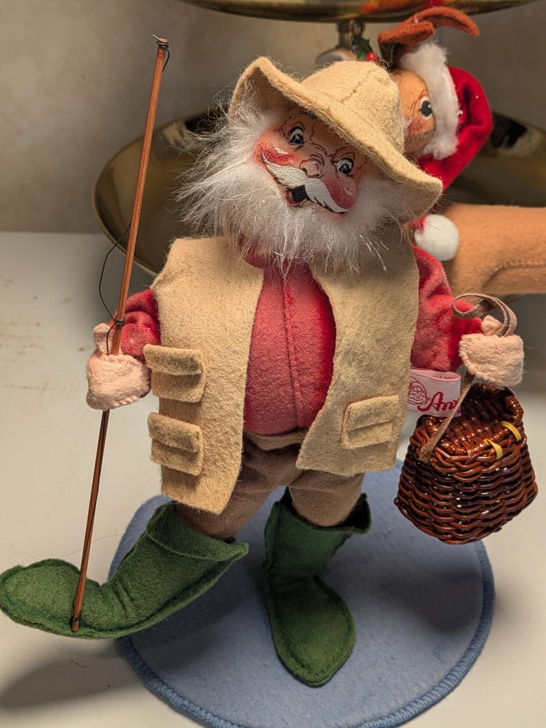 May include: A figurine of a bearded figure wearing a tan hat, a red shirt, a tan vest, and green boots. The figure holds a thin brown fishing rod in one hand and a small brown woven basket in the other. A white tag with red text is attached to the basket.