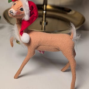 May include: A tan felt deer figurine wearing a red Santa hat with white trim and a white pom-pom. The deer has a white faux fur chest and tail, and a small sprig of green holly leaves with red berries attached to the top of the hat.