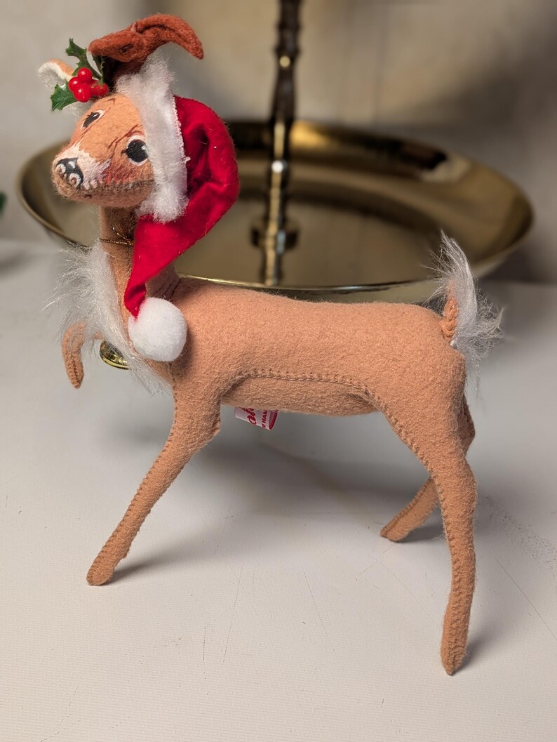 May include: A tan felt deer figurine wearing a red Santa hat with white trim and a white pom-pom. The deer has a white faux fur chest and tail, and a small sprig of green holly leaves with red berries attached to the top of the hat.