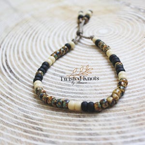 May include: A beaded bracelet featuring a mix of black, cream, and speckled beads. The bracelet is strung on a neutral-colored cord and includes the text "Twisted Knots by Sharon". The beads have a matte finish and are arranged in a repeating pattern.