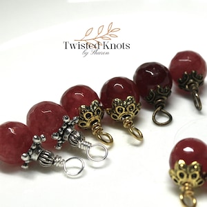 May include: Close-up of red gemstone beads with decorative metal caps. The caps have intricate designs in gold and silver tones. The beads are arranged on a white surface, with the text "Twisted Knots by Sharon" in the background.