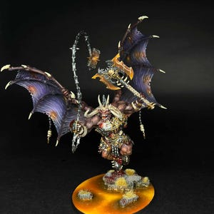 Puede incluir: Figura en miniatura detallada de un demonio alado con una expresión amenazante. La figura está pintada con una combinación de morados oscuros, naranjas y dorados. Empuña un hacha grande y una cadena con púas, de pie sobre una base texturizada.