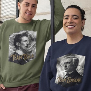 Könnte beinhalten: Zwei Personen tragen Sweatshirts mit Bildern von Nikola Tesla und Thomas Edison. Das Sweatshirt auf der linken Seite ist olivgrün mit einem Schwarzweißbild von Tesla und dem Text "Team Tesla". Das Sweatshirt auf der rechten Seite ist marineblau mit einem Schwarzweißbild von Edison und dem Text "Team Edison".