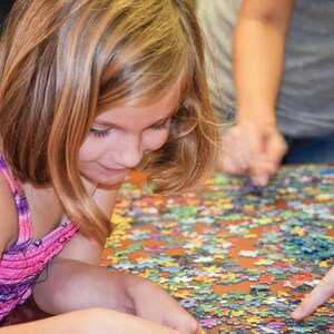May include: A young girl with long blonde hair is concentrating on a colorful jigsaw puzzle. She is wearing a pink and purple tie-dye tank top and is sitting at a table with the puzzle spread out in front of her.