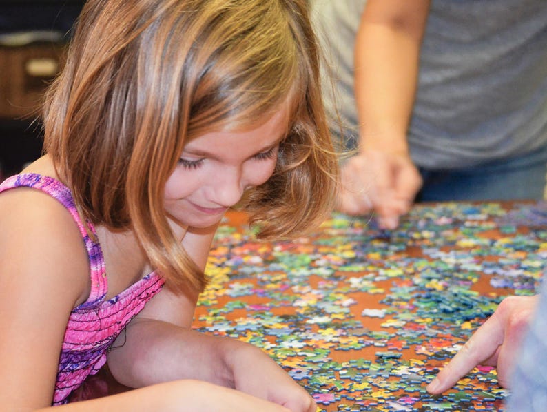 May include: A young girl with long blonde hair is concentrating on a colorful jigsaw puzzle. She is wearing a pink and purple tie-dye tank top and is sitting at a table with the puzzle spread out in front of her.