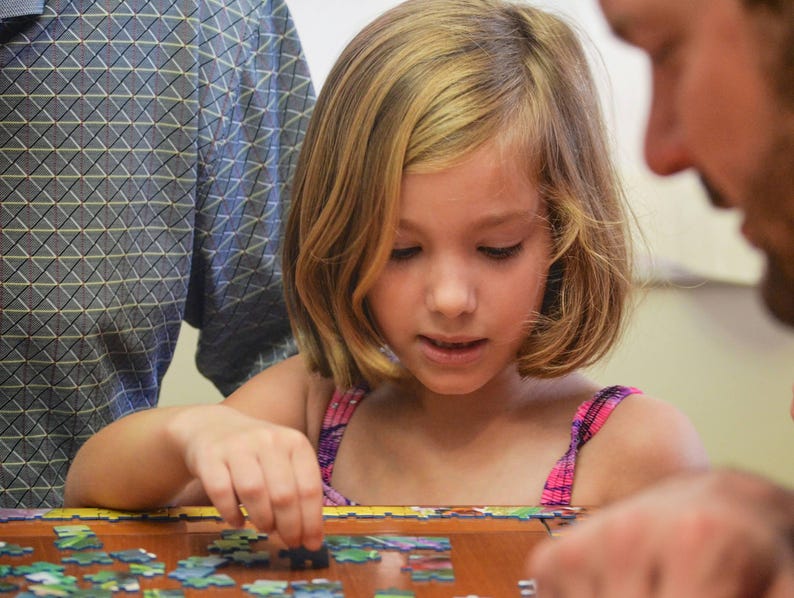 May include: A young girl with blonde hair is concentrating on putting together a jigsaw puzzle. She is wearing a pink and white tank top and is sitting at a table with a wooden surface.