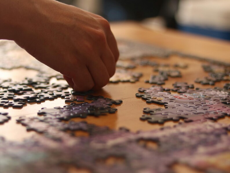 May include: A hand placing a puzzle piece onto a partially completed jigsaw puzzle. The puzzle pieces are various shades of purple and gray, with a wooden surface in the background.