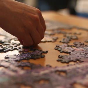 May include: A close-up of a person's hand placing a puzzle piece into a partially completed jigsaw puzzle. The puzzle pieces are purple and white with a floral pattern.