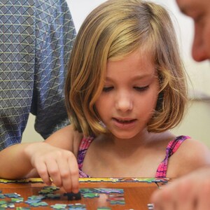 May include: A young girl with blonde hair and a pink and purple patterned top is focused on assembling a jigsaw puzzle. An adult is partially visible on the right side of the frame. Another adult is partially visible on the left side of the frame, wearing a blue patterned shirt.