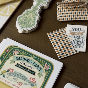 May include: A collection of stationery items featuring a sardine can label, decorative fork stickers, and patterned envelopes. The sardine label reads "Sardines Rares - Les Jolies - Made Different". A card reads "You are not like the others".