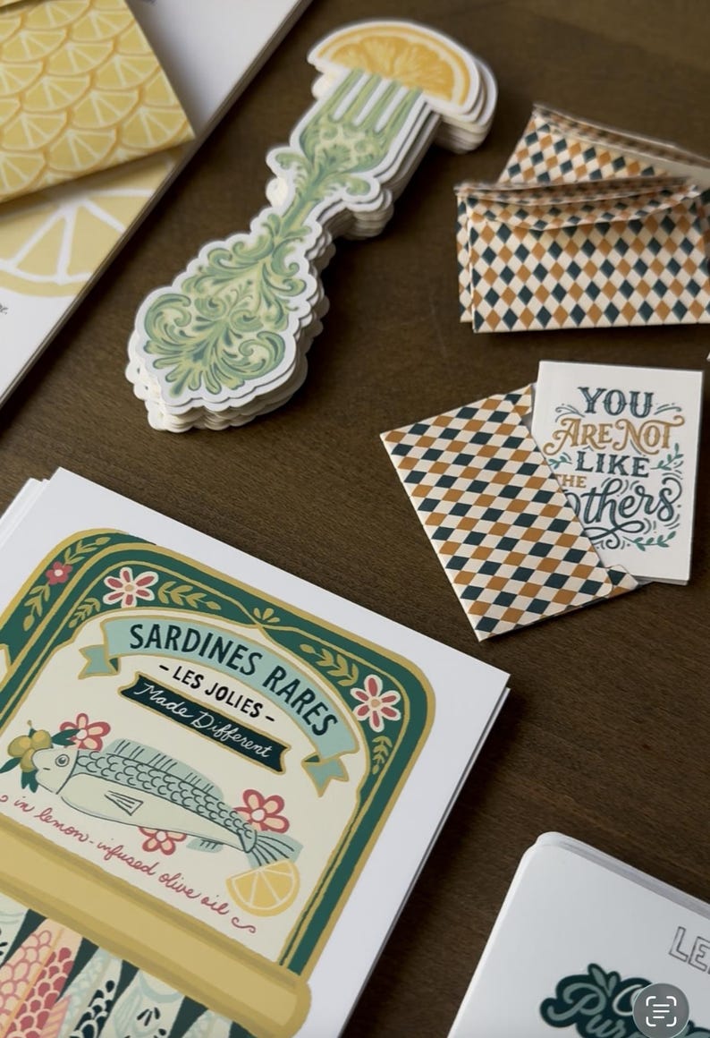 May include: A collection of stationery items featuring a sardine can label, decorative fork stickers, and patterned envelopes. The sardine label reads "Sardines Rares - Les Jolies - Made Different". A card reads "You are not like the others".
