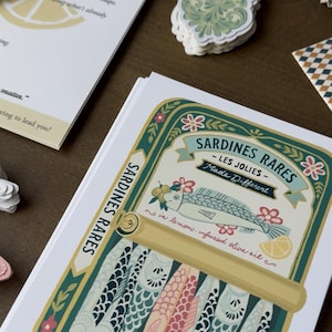 May include: A stack of paper goods featuring a sardine can illustration with the text "Sardines Rares - Les Jolies - Made Different". The can design includes fish, flowers, and lemon slices. Other items include a lemon-patterned card and decorative stickers.