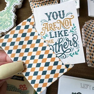 May include: A collection of stationery items, including cards and envelopes. The cards feature the text "YOU ARE NOT LIKE THE OTHERS" in teal and gold lettering. The envelopes have a repeating diamond pattern in teal, orange, and cream.