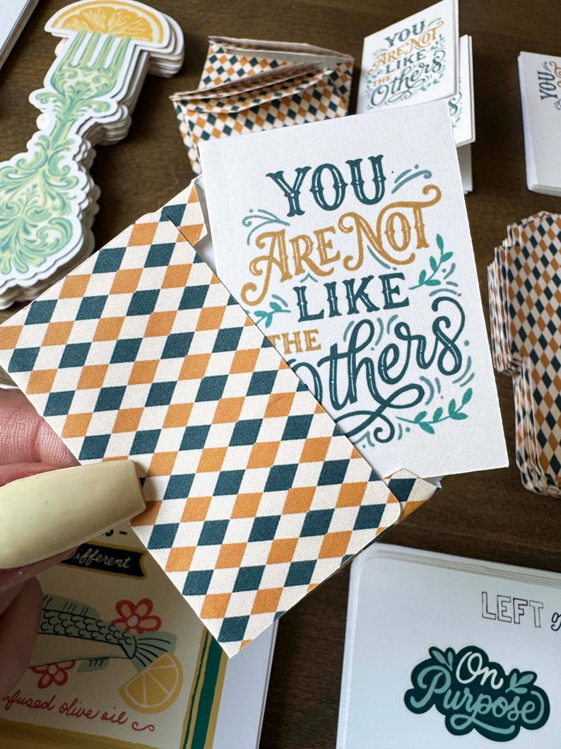 May include: A collection of stationery items, including cards and envelopes. The cards feature the text "YOU ARE NOT LIKE THE OTHERS" in teal and gold lettering. The envelopes have a repeating diamond pattern in teal, orange, and cream.