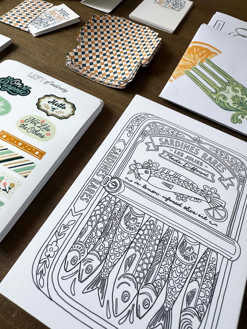May include: A collection of stationery items, including stickers, cards, and patterned paper. The stickers feature phrases like "On Purpose" and "Not Like the Others." A black and white illustration of sardines in a can is also visible, with the text "Sardines Rares."